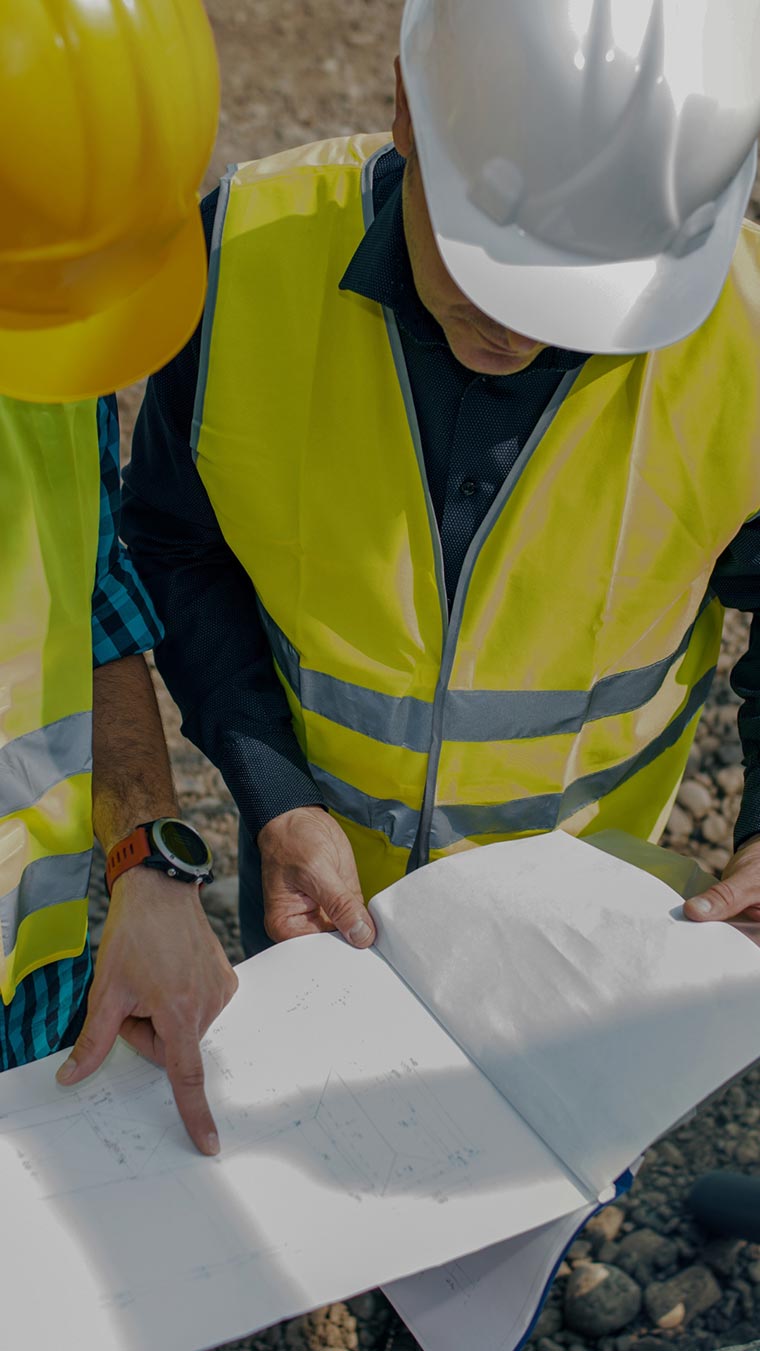 Deux professionnels du bâtiment, un homme et une femme, en tenue de chantier, examinant attentivement des plans et des documents papier sur un chantier de construction en extérieur avec des structures en arrière-plan