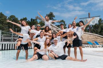 Photo de groupe des animateurs du camping Le Vieux Port saison 2025, posant souriants et en tenue décontractée au parc aquatique, entourés de structures aquatiques colorées, toboggans et bassins, sous un ciel clair d’été.