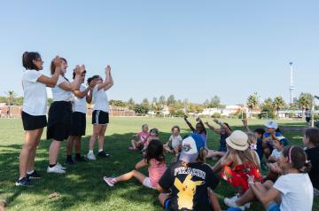 Animateurs du mini club donnant des consignes aux enfants rassemblés sur un terrain de football ombragé, dans une atmosphère calme et attentive, avant le début de la prochaine activité sportive, avec arbres fournissant de l’ombre et équipement sportif visible en arrière-plan.