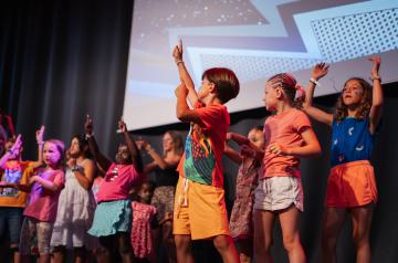 Spectacle hebdomadaire des enfants au camping Le Vieux Port, tous les vendredis soir dans la salle l'ARENA, mettant en vedette les jeunes stars des familles qui montent sur scène pour danser et jouer devant un public joyeux de parents et vacanciers, dans une atmosphère festive et chaleureuse.