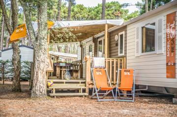 Mobil home moderne vu de près au camping Le Vieux Port dans les Landes, avec une grande terrasse en bois équipée de chaises longues confortables, entouré de pins majestueux typiques de la pinède landaise, offrant un cadre paisible et ombragé à quelques pas de la plage en bord de mer.