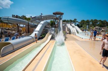 Sorties de plusieurs toboggans aquatiques au parc aquatique XXL du camping Le Vieux Port dans les Landes, avec des couloirs d'eau clairs dévalés par les vacanciers, entourés de structures colorées comme le Kamikaze, le Black Hole et le Pentagliss, dans un espace vaste et thématique inspiré de la piraterie, sous un ciel bleu et une végétation luxuriante typique de la pinède landaise.