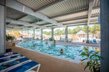 Grande piscine couverte et chauffée à 28°C du parc aquatique du camping 5 étoiles Le Vieux Port dans les Landes, entourée d’une structure moderne avec de larges ouvertures laissant entrer la lumière naturelle et facilitant les accès rapides, offrant un espace de baignade calme et convivial adapté à tous les âges, avec des zones de détente et des équipements de qualité pour profiter de la baignade en toute saison.