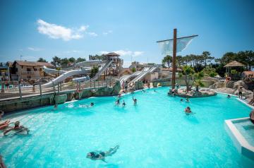 Grande piscine en forme de bateau située sur la plage du parc aquatique XXL du camping Le Vieux Port dans les Landes, avec un jacuzzi chauffé adjacent, entourée de sable fin et palmiers, offrant un cadre idyllique pour la détente au bord de l’océan Atlantique, sous un ciel bleu lumineux et une ambiance paradisiaque.