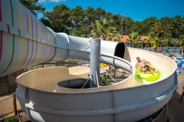 Un papa et son fils souriants partageant un moment de joie sur une bouée double en descente dans le toboggan à virages du parc aquatique XXL du camping Le Vieux Port dans les Landes, entourés d’eaux claires, glissant à toute vitesse à travers les courbes du toboggan dans une ambiance familiale et ludique.