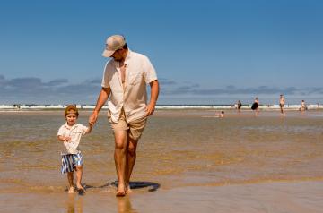 Un père et son fils debout sur le littoral landais, les pieds dans l'eau claire et calme à marée basse, entourés d’une étendue de sable fin et de petites baïnes, sous un ciel dégagé, avec l’horizon maritime à perte de vue, dans un cadre naturel typique de la côte landaise marqué par la lumière douce, la sérénité et la beauté du bord de mer.