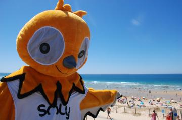 Soly, la mascotte joyeuse du camping Le Vieux Port dans les Landes, posant avec les vacanciers souriants sur la plage de sable fin aux eaux turquoise de l’océan Atlantique, sous un ciel lumineux, incarnant la convivialité et le bonheur d’un séjour inoubliable en bord de mer dans un cadre naturel préservé et familial.