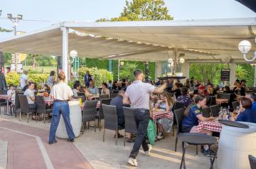 Serveurs en tenue professionnelle servant des boissons fraîches et des apéritifs à des vacanciers souriants assis aux tables en terrasse du bar Le VP, au camping Le Vieux Port dans les Landes. L’ambiance est conviviale et détendue, avec des clients profitant du soleil sur une grande terrasse en bois, entourée de pins, dans un cadre chaleureux et festif en bord de mer.