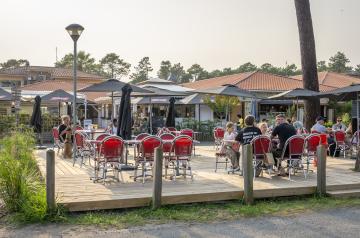 Nouvelle terrasse spacieuse et moderne du bar Le VP au camping Le Vieux Port dans les Landes, avec des tables en bois clair et chaises confortables disposées sous une pergola offrant de l’ombre, entourée de plantes et de lumières d’ambiance, créant un espace convivial et élégant où les vacanciers peuvent se détendre, savourer un verre ou un repas en profitant de la douceur du climat landais.