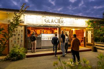 Glacier du camping Le Vieux Port ouvert la nuit, éclairé par des lumières chaleureuses qui attirent les vacanciers désireux de savourer des glaces artisanales et des desserts sucrés. Le stand est animé par un personnel souriant, avec une ambiance festive et conviviale sous un ciel étoilé, offrant un moment de détente gourmand en fin de journée dans ce camping 5 étoiles en bord de l’océan Atlantique.