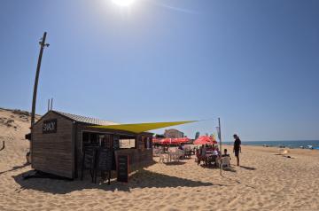 Snack bar situé directement sur la plage, les pieds dans le sable, au camping Le Vieux Port dans les Landes, accueillant des vacanciers assis à des tables, profitant de boissons fraîches et de snacks savoureux dans une atmosphère conviviale et détendue au bord de l’océan Atlantique. L’espace est entouré de la dune, avec vue sur la plage et accès direct par un chemin sécurisé depuis le camping.