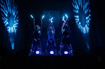 Spectacle dynamique à l’Arena du camping Le Vieux Port avec trois danseurs évoluant sur une scène éclairée, portant des costumes futuristes et manipulant des accessoires LED colorés qui créent des jeux de lumière hypnotiques. La scène spacieuse et équipée de matériel professionnel met en valeur la chorégraphie synchronisée et l’énergie des artistes, dans une ambiance moderne et immersive, captivant un public nombreux dans la plus grande salle de spectacles des Landes, offrant une belle expérience visuelle.