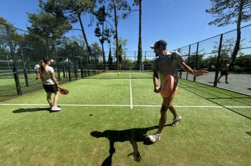 Des joueurs disputant un match de padel sur un terrain clôturé de verre et grillage, sous un ciel dégagé. Quatre participants en tenues de sport, équipés de raquettes spécifiques au padel, sont en pleine action, se concentrant sur la balle qui vole entre eux. Le terrain, doté d'un sol en gazon artificiel, est entouré d'un public limité ou d'un cadre naturel, avec une ambiance sportive et conviviale, où l'agilité, la rapidité et la stratégie sont essentielles au jeu.