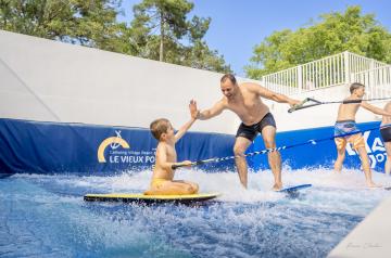 Un père et son jeune fils se tapant la main avec enthousiasme, debout dans l’eau peu profonde du bassin de surf à la Vieux Port Surfing Wave, dans le parc aquatique. Le père porte un maillot de bain coloré et un sourire large, tandis que le fils, également en maillot de bain, montre une expression de joie et de concentration. En arrière-plan, on voit la grande machine à vagues et d’autres activités aquatiques, sous un ciel bleu clair et un soleil brillant. La scène respire la complicité, le plaisir.