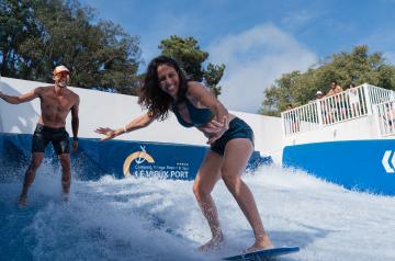 Une femme participante à l’animation surfing contest adultes sur la Vieux Port Surfing Wave du parc aquatique du camping Le Vieux Port. Elle est debout en équilibre sur sa planche de surf artificielle, concentrée et souriante, vêtue d’une combinaison de sport adaptée. Autour d’elle, l’eau en mouvement simule des vagues dynamiques créées par la machine à surf WaveMotion, dans un environnement sécurisé et moderne. En arrière-plan, on distingue d’autres participants et animateurs encadrant l’activité.