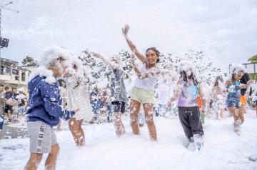 Pré-adolescente souriante, cheveux mouillés, au milieu d'une soirée mousse animée, entourée de bulles blanches et d'une foule joyeuse. Elle porte un t-shirt léger humide, dansant et s'amusant dans une ambiance festive, colorée par les jeux de lumière et la mousse abondante qui forme un nuage dense autour des participants.