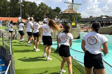 Animateurs dynamiques en pleine chorégraphie de danse du midi au camping, situés dans un parc aquatique ensoleillé. Ils portent des tenues colorées et légères adaptées à l’ambiance estivale, avec en arrière-plan des toboggans et bassins d’eau. L’atmosphère est joyeuse et festive, rassemblant vacanciers de tous âges qui participent activement aux mouvements rythmés et synchronisés, symbolisant l’esprit convivial et énergique des animations de camping.