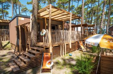 Vue extérieure d'un locatif moderne et élégant avec façade en bois chaleureux, grandes baies vitrées ouvertes sur un jardin verdoyant, terrasse en bois avec mobilier design pour profiter des beaux jours, ciel bleu lumineux en arrière-plan, ambiance accueillante et confortable invitant à la découverte et à la détente.