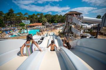 Trois enfants heureux descendant en même temps un grand toboggan coloré dans un parc de jeux en plein air, leurs visages rayonnants de joie et d’excitation alors qu’ils glissent rapidement vers le bas sous un ciel ensoleillé