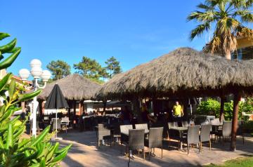 Photo du restaurant La Trattoria avec ses grandes paillotes en toit de chaume, tables et chaises en bois disposées dessous à l'ombre, ambiance conviviale et ombragée, lumière naturelle filtrée par la végétation, décor rustique et chaleureux invitant à la détente et au repas en plein air.