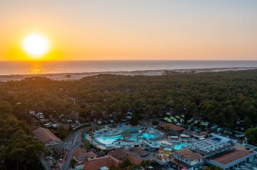 Photo aérienne prise par drone au coucher du soleil, avec vue panoramique sur l'horizon où le soleil se couche sur l'océan Atlantique, lumière dorée reflétée sur les vagues, camping visible en bas de l'image, entouré de verdure, avec son grand parc aquatique remarquable comprenant piscines et toboggans, atmosphère paisible et chaleureuse typique des heures dorées.