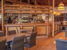 Intérieur chaleureux du bar bistro de la brasserie du meilleur camping du Sud-Ouest des Landes, avec un comptoir en bois verni, des tables conviviales joliment dressées, des chaises confortables, une décoration authentique mêlant touches modernes et esprit landais, de grandes baies vitrées laissant entrer la lumière naturelle, un éclairage doux mettant en valeur les bouteilles et verres derrière le bar, et une ambiance accueillante idéale pour partager un verre ou un repas en vacances.