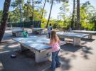 Jeunes personnes et adolescents en train de jouer au ping-pong sur une terrasse extérieure au camping Le Vieux Port, avec des tables de ping-pong en bois, entourés de végétation verdoyante et de structures de camping, ambiance conviviale et ensoleillée