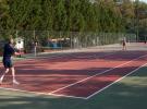 Groupe de trois hommes et une femme en tenue de sport, engagés dans un match de tennis sur un court en plein air au camping, avec des arbres et des tentes en arrière-plan, profitant de leurs vacances ensoleillées, montrant leur enthousiasme et leur concentration lors de la partie