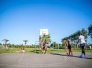 Jeunes ados et jeunes adultes jouant au basketball lors d'une rencontre sportive au camping Le Vieux Port à Messanges, où un joueur est en train de marquer un panier avec un saut spectaculaire, sous un ciel ensoleillé, entourés de spectateurs enthousiastes et de paysages naturels aquitains