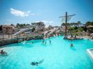 Grande piscine en forme de bateau située sur la plage du parc aquatique XXL du camping Le Vieux Port dans les Landes, avec un jacuzzi chauffé adjacent, entourée de sable fin et palmiers, offrant un cadre idyllique pour la détente au bord de l’océan Atlantique, sous un ciel bleu lumineux et une ambiance paradisiaque.