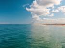 Vue depuis l’océan vers la plage de Messanges, avec sur la moitié gauche de l’image l’océan Atlantique s’étendant à perte de vue sous un ciel totalement dégagé sans nuages, et sur la moitié droite le phare de Messanges se détachant près du rivage sous un ciel partiellement couvert de nuages plus sombres au-dessus de la côte.