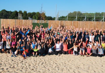 Groupe d'environ cent personnes réunies en extérieur lors d'une activité de team-building, levant toutes les mains en l'air en signe de participation et d'enthousiasme, avec des sourires, des tenues décontractées