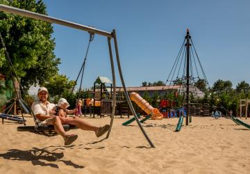 Un papa tenant tendrement son bébé dans ses bras à l'aire de jeux du camping, tous deux pieds nus dans le sable, profitant d'un moment de complicité et de détente en plein air