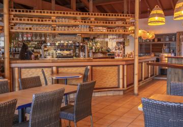 Intérieur chaleureux du bar bistro de la brasserie du meilleur camping du Sud-Ouest des Landes, avec un comptoir en bois verni, des tables conviviales joliment dressées, des chaises confortables, une décoration authentique mêlant touches modernes et esprit landais, de grandes baies vitrées laissant entrer la lumière naturelle, un éclairage doux mettant en valeur les bouteilles et verres derrière le bar, et une ambiance accueillante idéale pour partager un verre ou un repas en vacances.