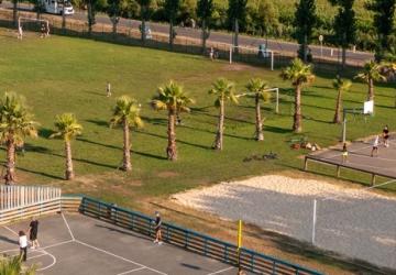 Vue panoramique de la plaine des sports du camping Le Vieux Port, avec terrains de football et de volley-ball en herbe, entourés d'arbres verts et de zones de détente, offrant un espace convivial pour les activités sportives et récréatives en plein air