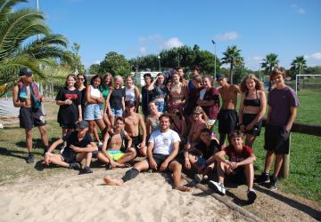 Photo de groupe montrant une dizaine d'adolescents souriants et détendus, posant ensemble sur la terrasse de la plaine des sports au camping, avec des arbres et des installations sportives en arrière-plan, lors d'une activité ou d'une sortie organisée au sein du club ados