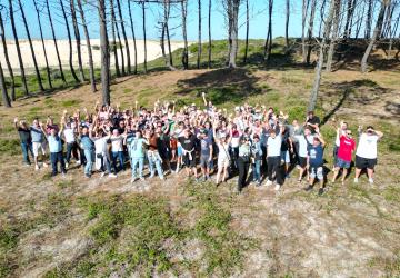 Vue aérienne réalisée par drone d’un groupe d’environ 100 participants réunis lors d’un événement de teambuilding. Tous sont rassemblés derrière une dune de sable, en extérieur, avec les bras levés vers le ciel dans une ambiance festive et conviviale, exprimant la cohésion d’équipe et l’enthousiasme collectif. La prise de vue met en valeur la composition du groupe, l’environnement naturel et le dynamisme du moment.