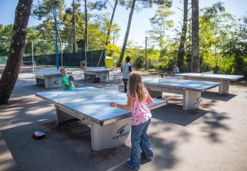 Jeunes personnes et adolescents en train de jouer au ping-pong sur une terrasse extérieure au camping Le Vieux Port, avec des tables de ping-pong en bois, entourés de végétation verdoyante et de structures de camping, ambiance conviviale et ensoleillée