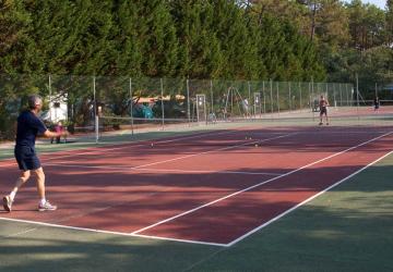 Groupe de trois hommes et une femme en tenue de sport, engagés dans un match de tennis sur un court en plein air au camping, avec des arbres et des tentes en arrière-plan, profitant de leurs vacances ensoleillées, montrant leur enthousiasme et leur concentration lors de la partie