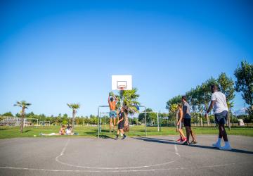Jeunes ados et jeunes adultes jouant au basketball lors d'une rencontre sportive au camping Le Vieux Port à Messanges, où un joueur est en train de marquer un panier avec un saut spectaculaire, sous un ciel ensoleillé, entourés de spectateurs enthousiastes et de paysages naturels aquitains