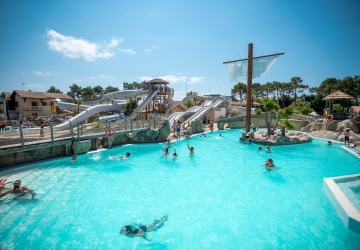 Grande piscine en forme de bateau située sur la plage du parc aquatique XXL du camping Le Vieux Port dans les Landes, avec un jacuzzi chauffé adjacent, entourée de sable fin et palmiers, offrant un cadre idyllique pour la détente au bord de l’océan Atlantique, sous un ciel bleu lumineux et une ambiance paradisiaque.