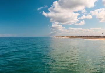 Vue depuis l’océan vers la plage de Messanges, avec sur la moitié gauche de l’image l’océan Atlantique s’étendant à perte de vue sous un ciel totalement dégagé sans nuages, et sur la moitié droite le phare de Messanges se détachant près du rivage sous un ciel partiellement couvert de nuages plus sombres au-dessus de la côte.