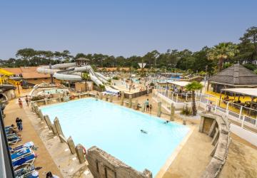 Vue panoramique en juin du parc aquatique XXL du camping 5 étoiles Le Vieux Port dans les Landes, avec plusieurs piscines extérieures chauffées, toboggans aquatiques géants, espace Aquasplash thématisé pirates avec crâne de pirate, pataugeoires pour enfants, jacuzzis, transats occupés par les vacanciers et grande pinède en arrière-plan sous un ciel bleu estival