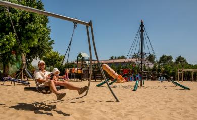 Un papa tenant tendrement son bébé dans ses bras à l'aire de jeux du camping, tous deux pieds nus dans le sable, profitant d'un moment de complicité et de détente en plein air