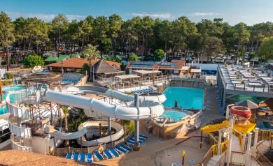 Vue panoramique sur le vaste parc aquatique du camping Le Vieux Port à Messanges, prise depuis le sommet des toboggans colorés de l’espace aquasplash. On distingue en contrebas les onze toboggans, dont le pentagliss géant, se jetant dans de grands bassins animés où enfants et familles s’amusent. À gauche, des pataugeoires ludiques et des jeux d’eau attirent les plus petits, tandis qu’au centre, le lagon à vagues chauffé accueille de nombreux baigneurs. Au second plan, des palmiers et la pinède de 30 hectare
