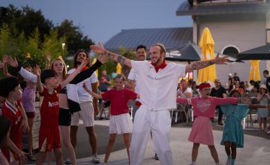 Soirée féria animée au camping Le Vieux Port dans les Landes en 2025, avec des vacanciers et animateurs portant des tenues traditionnelles colorées, dansant et chantant au rythme de musiques festives et entraînantes. L’ambiance est conviviale et chaleureuse, avec des guirlandes lumineuses, des décorations festives et une foule joyeuse rassemblée autour de l’ARENA, la grande salle de spectacles du camping, sous un ciel étoilé, pour une nuit mémorable de fête et de partage.