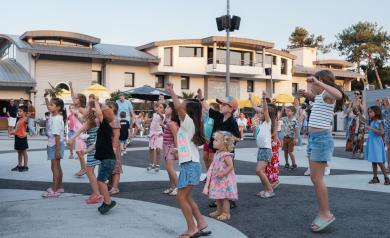 Soirée Soly Disco animée au camping Le Vieux Port dans les Landes en 2025, avec une foule d’enfants joyeux dansant au premier plan sous des jeux de lumières colorées et une ambiance musicale entraînante. La mascotte Soly est présente, entourée d’animateurs dynamiques, sur une piste de danse extérieure, créant une atmosphère festive, familiale et conviviale typique des animations pour enfants du camping 5 étoiles.