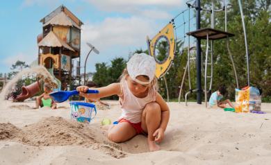 Enfant d'environ 7 ans souriant et jouant dans l'immense bac à sable de l'aire de jeux Kids du camping Le Vieux Port à Messanges, réservé aux 6-12 ans. Le garçon porte un t-shirt coloré et un short, ses pieds nus s’enfoncent dans le sable doux tandis qu’il grimpe sur le bateau bleu emblématique, entouré de balançoires, toboggans et portiques adaptés à son âge. Au fond, d'autres enfants s’amusent et découvrent les installations ludiques sous la surveillance de leurs parents, dans un cadre naturel