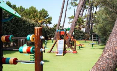 Aire de jeux pour enfants de 2 à 6 ans, installée sur un sol amortissant de couleur verte, comprenant toboggan et jeux à ressort, située au pied d’une dune en lisière de pinède sous les pins maritimes.