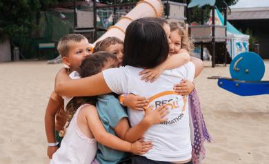 Animatrice souriante faisant un câlin collectif avec plusieurs enfants joyeux du mini club dont elle a la responsabilité, dans un cadre chaleureux et convivial.